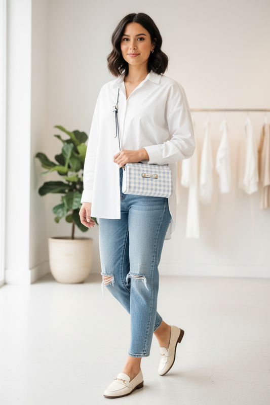 A woman models a white button-down shirt, light-wash distressed jeans, and cream loafers, carrying a small blue-and-white gingham handbag with a black strap in a bright, modern room.