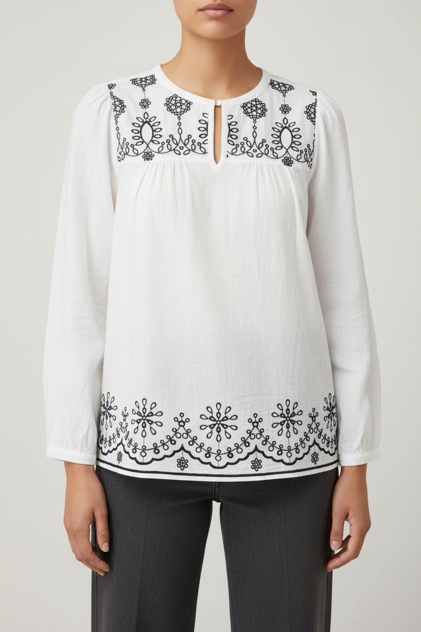 A woman wearing a white long-sleeve blouse with black floral embroidery around the neckline and hem, paired with dark gray trousers, against a light gray studio background.