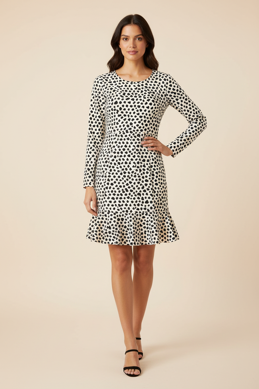 A woman modeling a long-sleeve knee-length dress with a black and white spotted pattern, standing against a solid beige studio background.