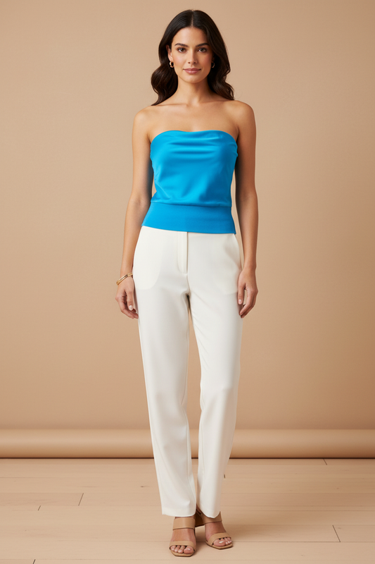 A woman modeling a bright blue strapless tube top paired with white straight-leg pants and beige heeled sandals, standing against a warm beige studio backdrop.