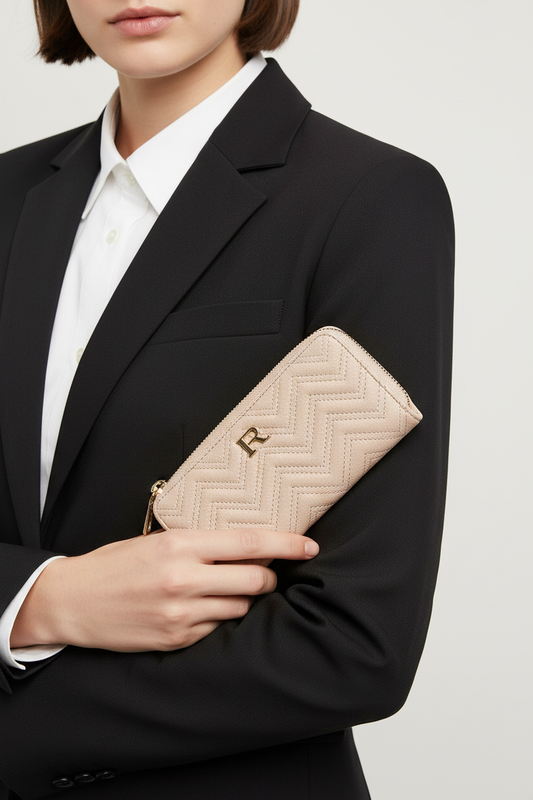 A close-up of a model in a black blazer and white collared shirt holding a beige chevron-patterned zip-around wallet with a gold logo clasp.