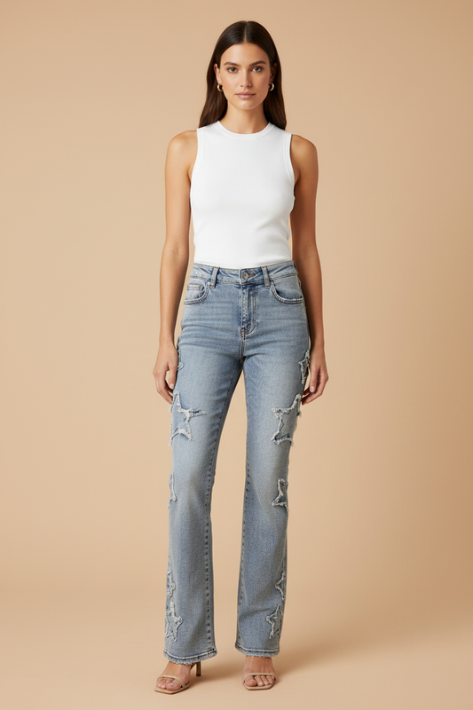 A woman modeling light-wash blue distressed jeans with a white sleeveless top, standing against a solid beige studio background.