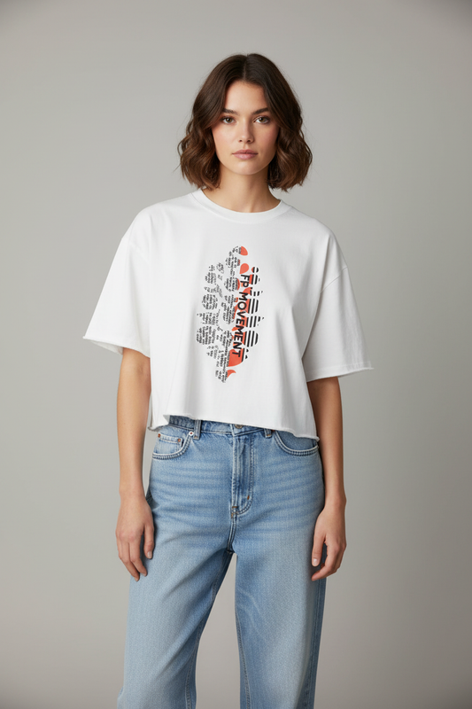 A woman modeling a white cropped t-shirt with a graphic print of the word 'BELIEVE' in red, black, and gray, paired with light blue jeans, against a neutral gray background.