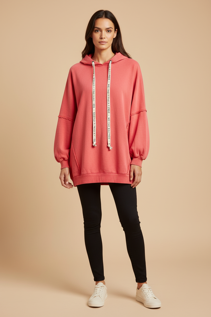 A woman modeling an oversized coral pink hoodie with white drawstrings featuring black text, paired with black skinny pants and white sneakers, against a solid beige studio background.