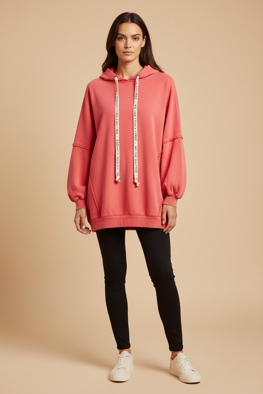 A woman modeling an oversized coral pink hoodie with white drawstrings featuring black text, paired with black skinny pants and white sneakers, against a solid beige studio background.