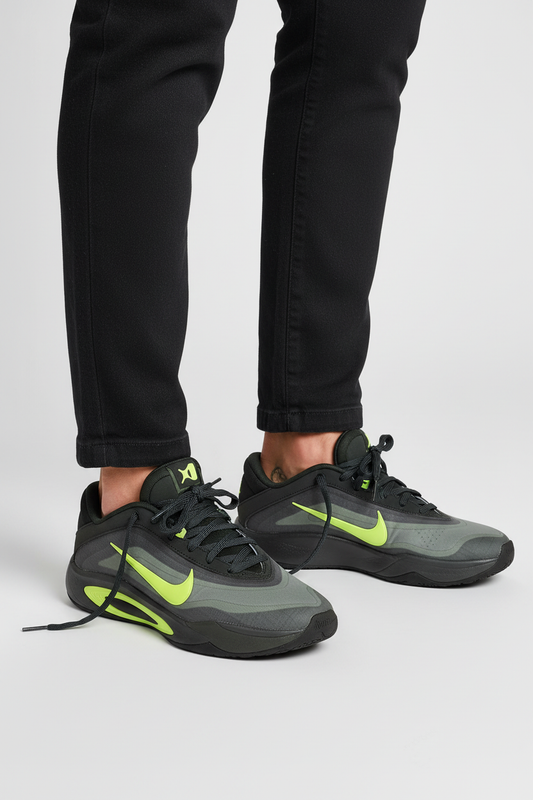 A pair of Nike low-top sneakers in dark gray and black with bright neon yellow-green accents, shown on a model wearing black pants against a light gray studio background.