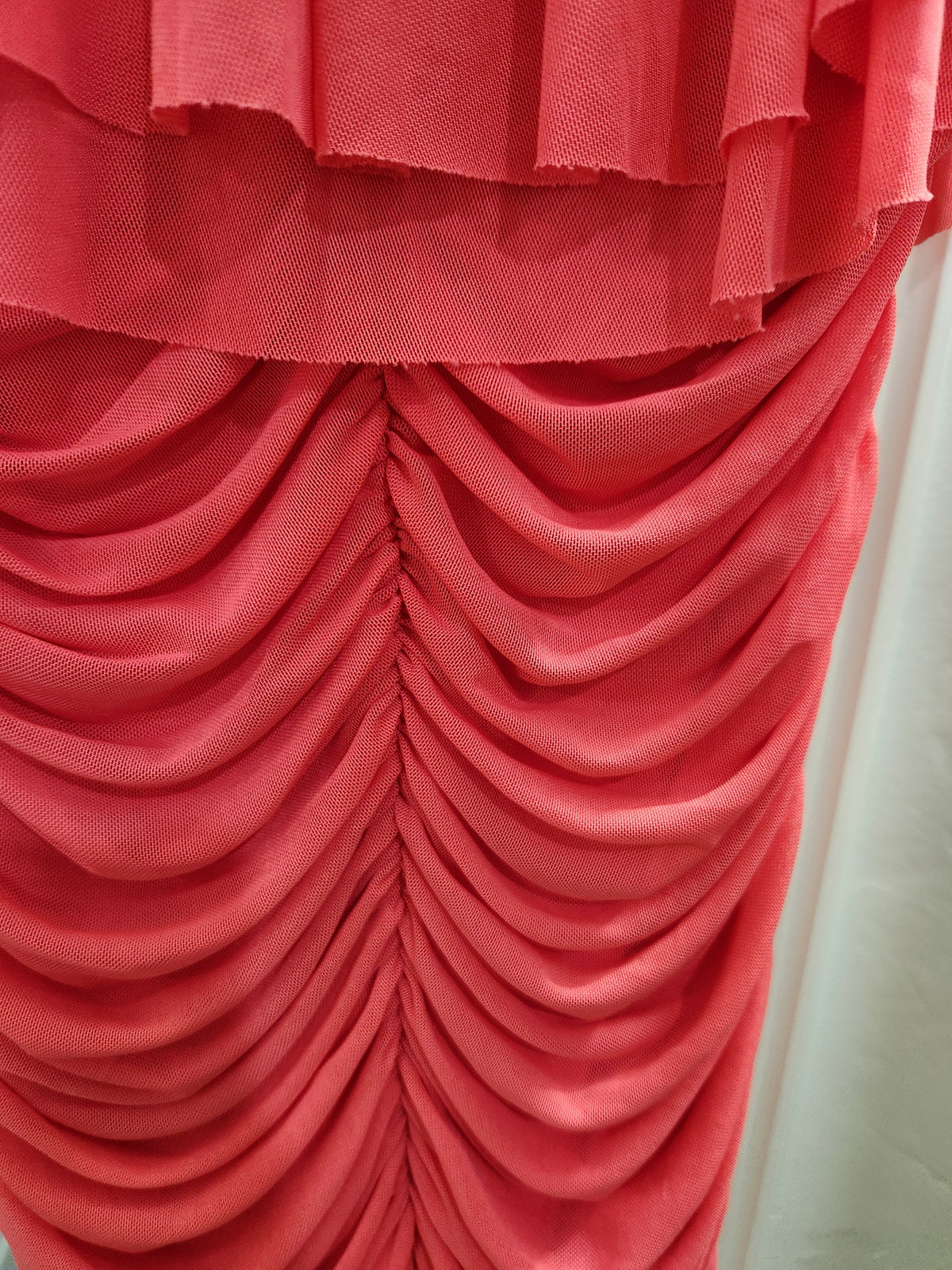 A close-up of a coral pink or salmon red ruched fabric garment, showing gathered pleats and a ruffled trim at the top, with a white fabric backdrop visible on the right side.
