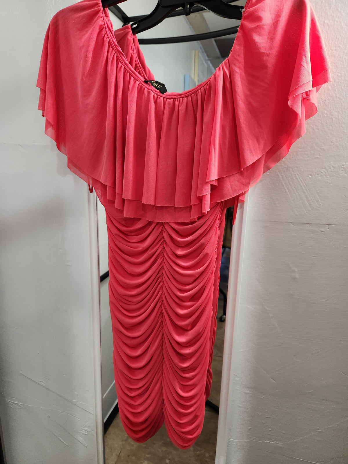 A bright pinkish-red off-the-shoulder dress with layered ruffle sleeves and a gathered ruched bodice, hanging on a black hanger against a white wall.