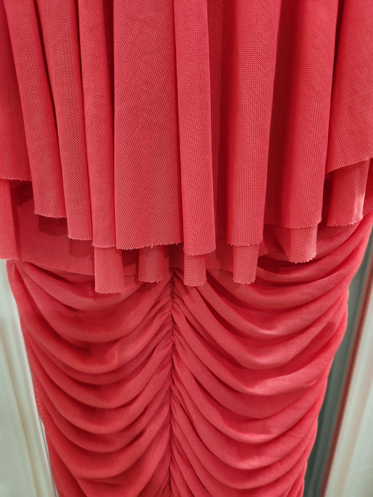 A close-up of a coral pink pleated fabric, showing both a fine textured knit top layer and a gathered, ruched velvet-like bottom layer.