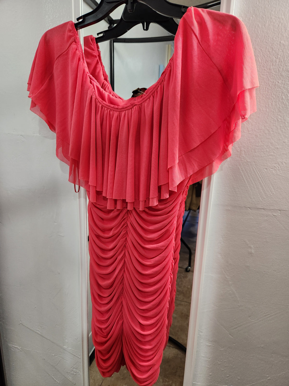 A bright coral pink draped dress with ruffled sleeves and a ruched skirt, hanging on a black hanger against a white wall.