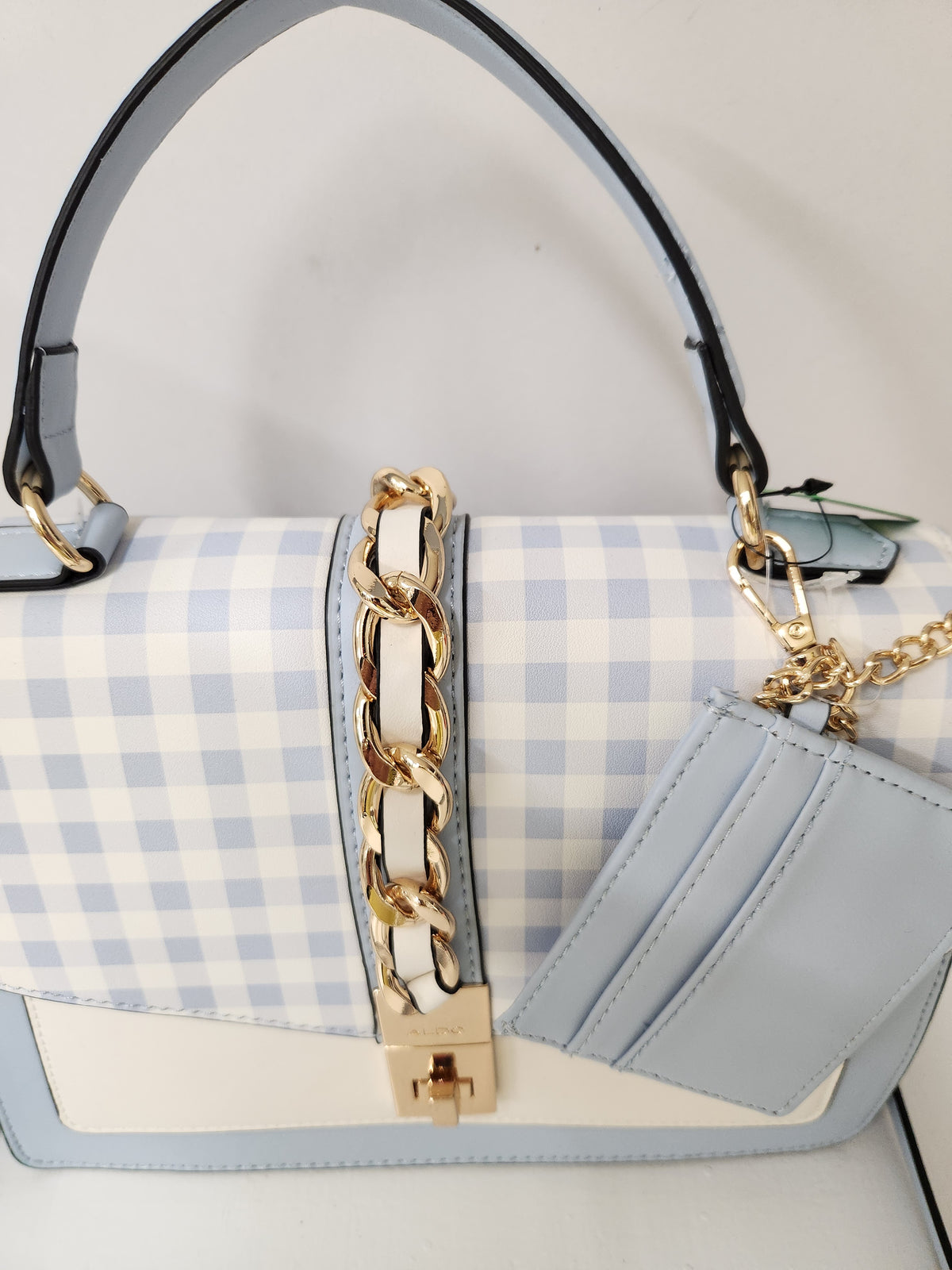 A light blue and white gingham handbag with a light blue leather front pocket, gold-tone chain strap, and gold clasp, shown against a plain light background.