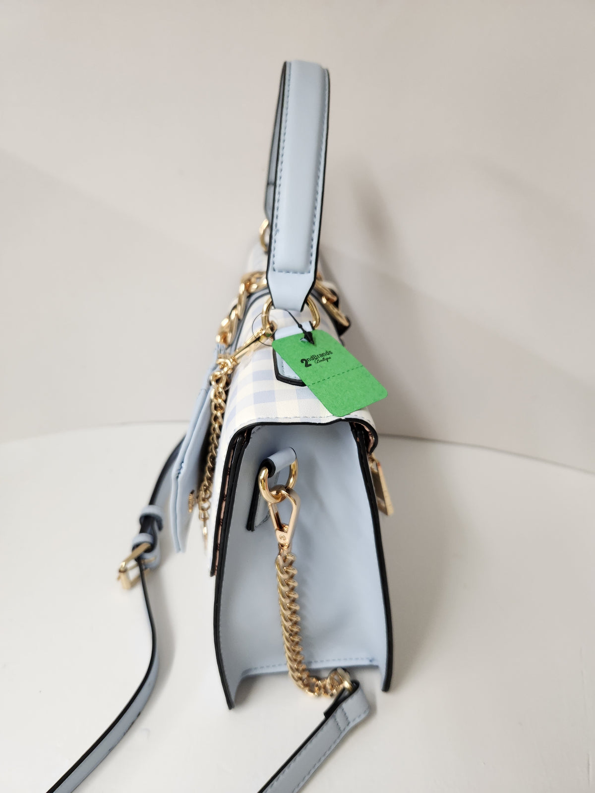 A light blue leather handbag with gold chain details, a white and blue striped fabric accent, and a green hangtag, photographed on a light gray background.