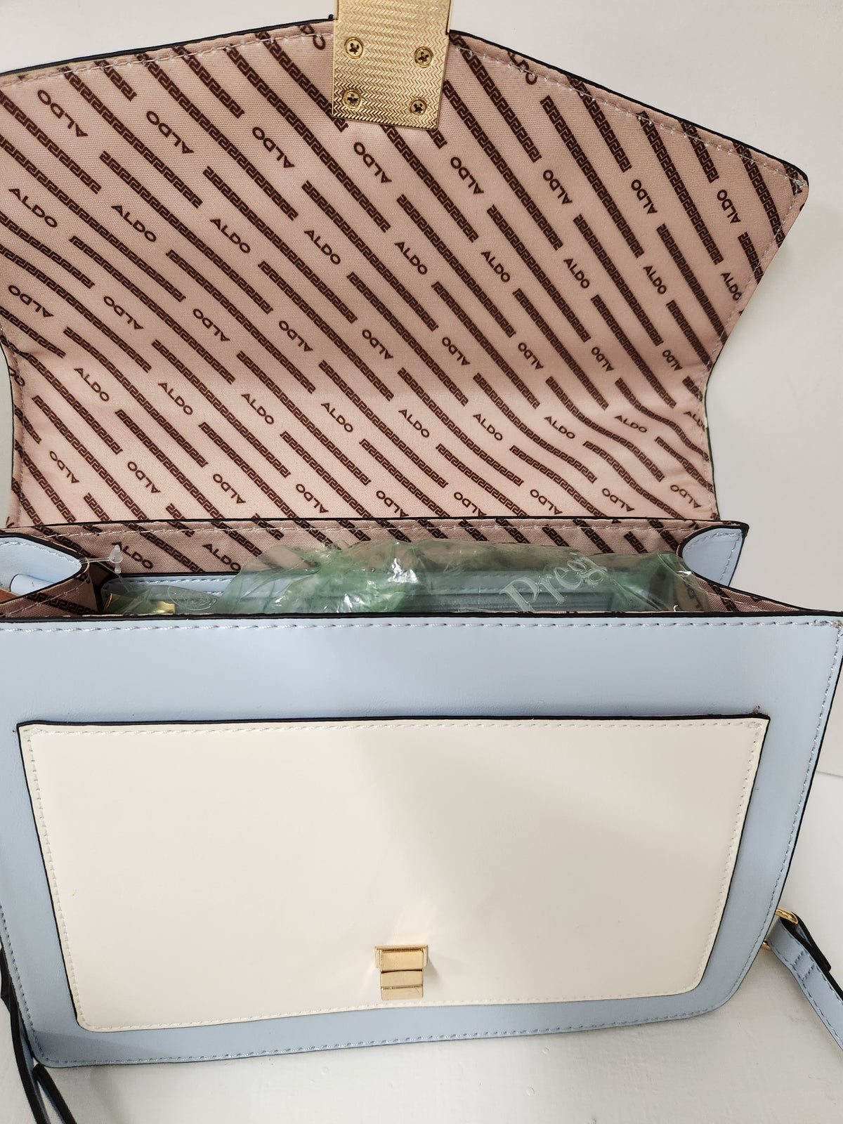 A light blue and off-white shoulder bag shown open, with a beige interior lined with repeating 'ALDO' branding. A green item is tucked inside, and the bag has a small gold-tone clasp on the front flap.