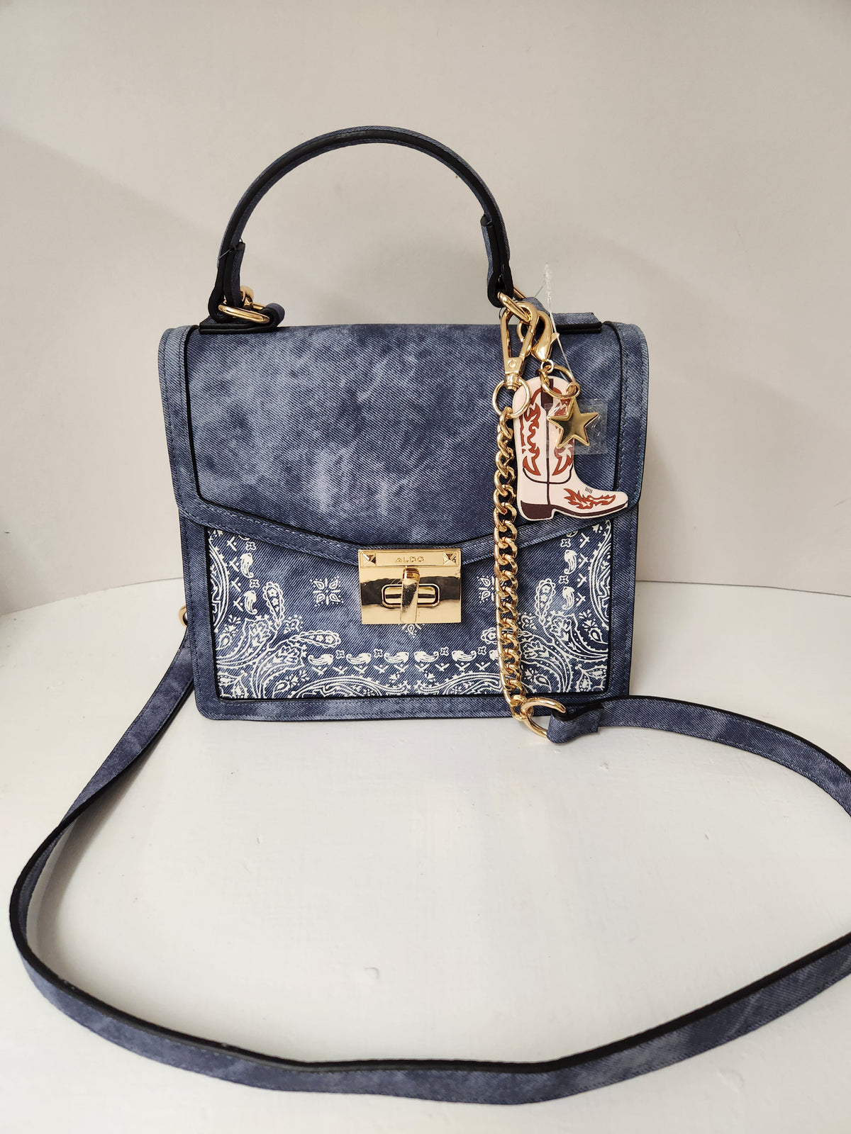 A blue suede handbag with a western-themed paisley pattern panel on the front, a gold-tone clasp, a black top handle, a gold chain strap, and a decorative boot-shaped charm.