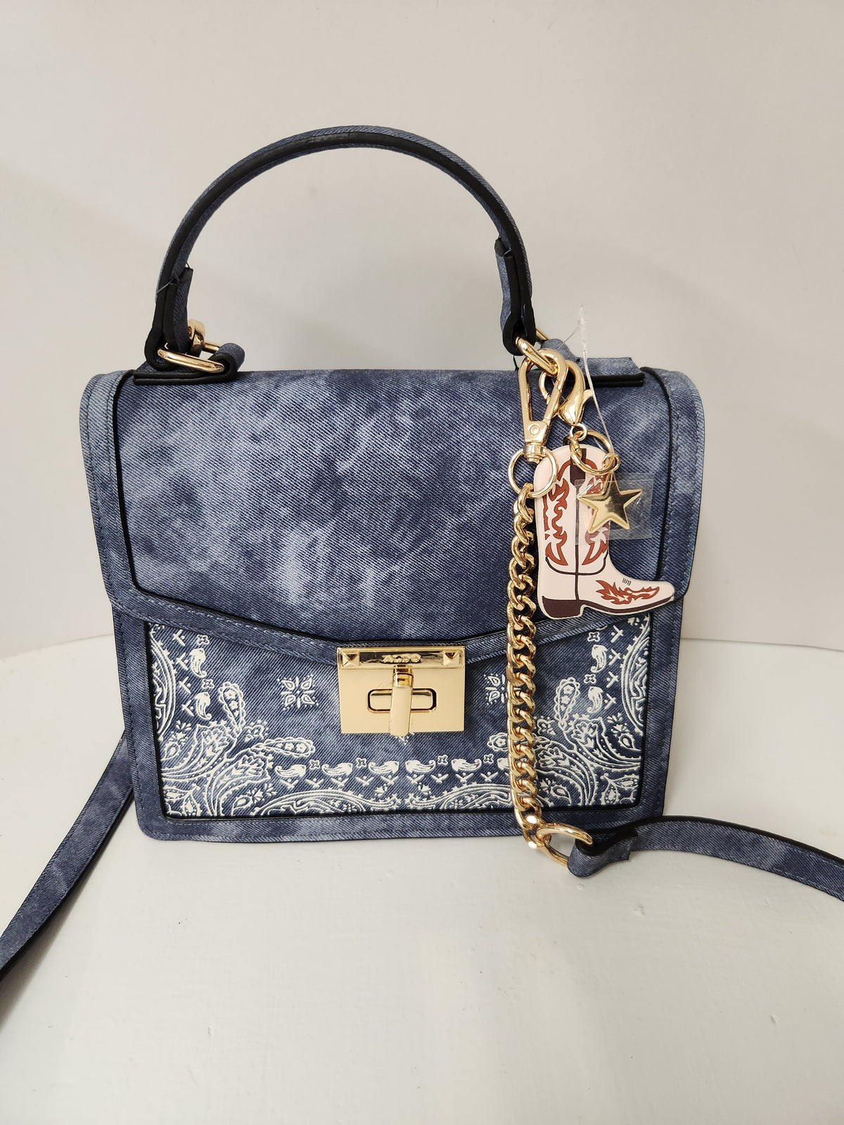 A blue suede handbag with a white paisley bandana print on the lower front flap, featuring a black top handle, gold-tone lock and chain, and a cowboy boot charm.