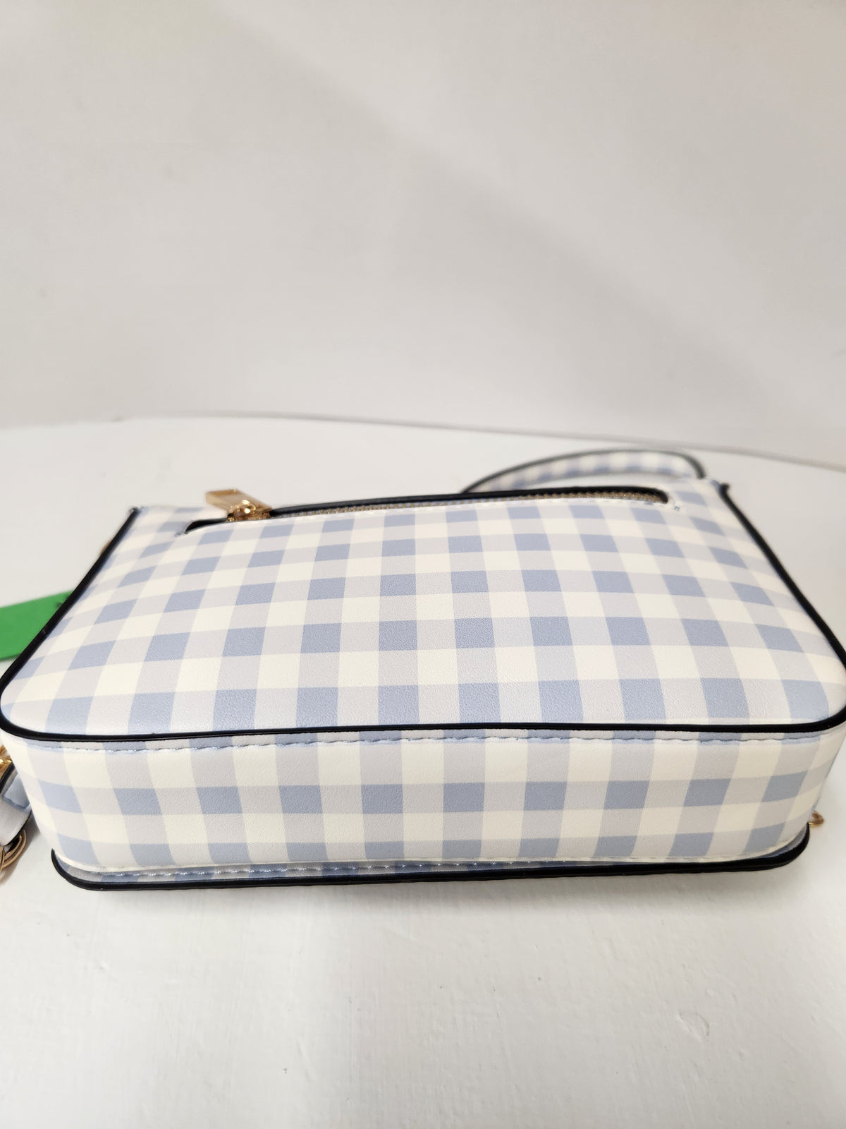 A small rectangular handbag with a light blue and white gingham check pattern, black piping, and a gold zipper, resting on a white surface against a white background.