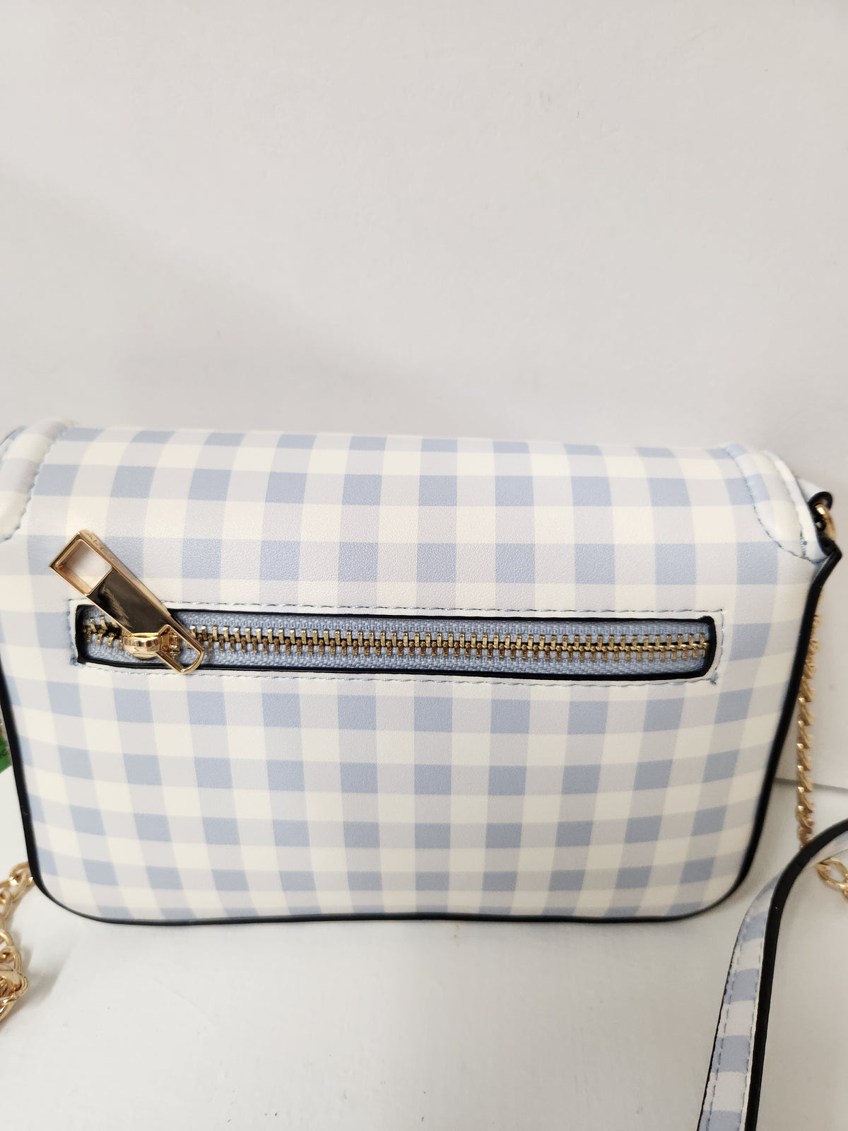 A small crossbody bag with a light blue and white gingham check pattern, featuring a horizontal zippered pocket with a gold-tone zipper and a gold-tone zipper pull. The bag has black trim and a gold chain strap, shown against a plain white background.