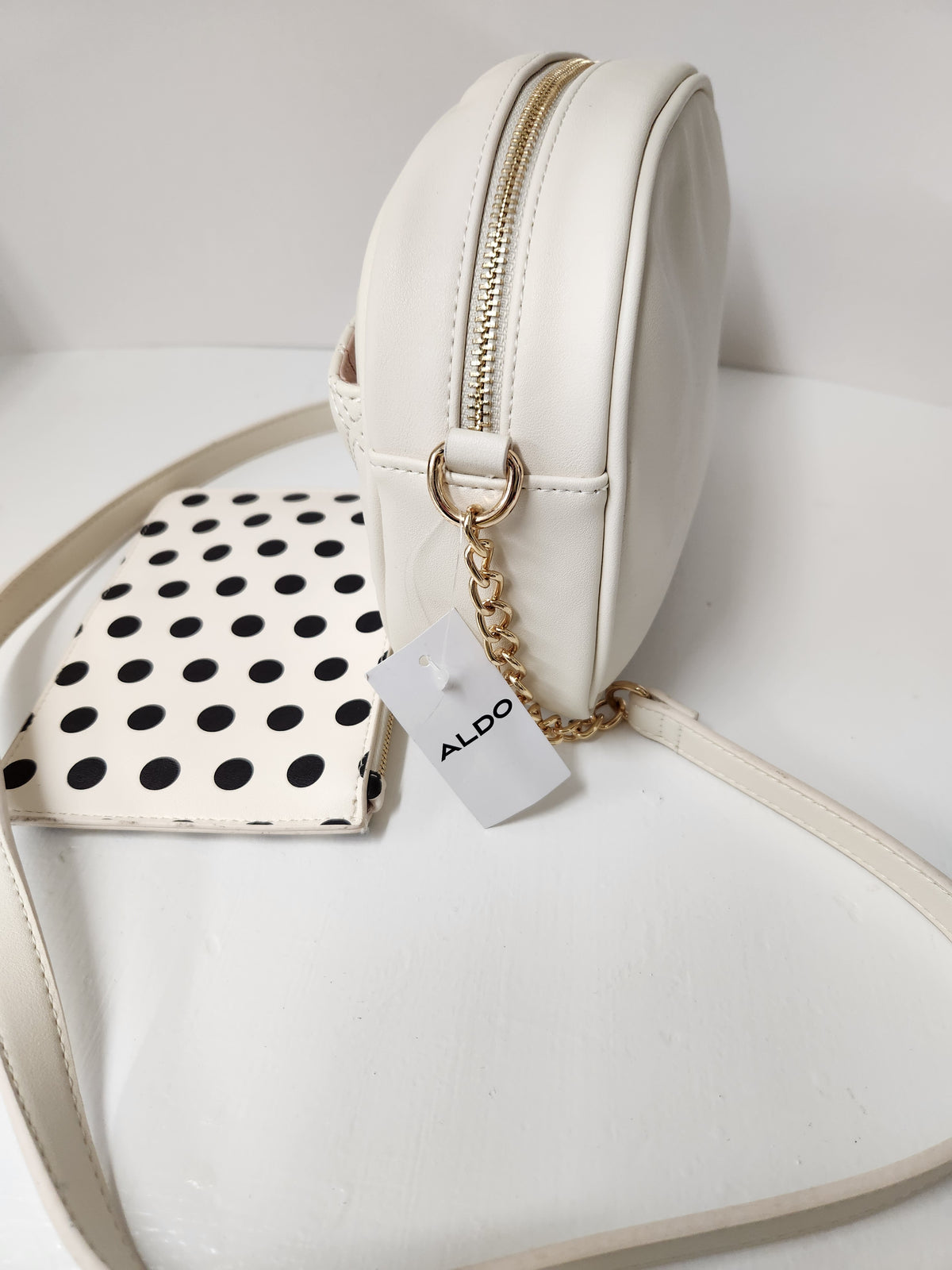 An off-white faux leather crossbody bag with a gold chain strap and a white ALDO brand tag, displayed next to a matching off-white and black polka dot pouch on a white surface.