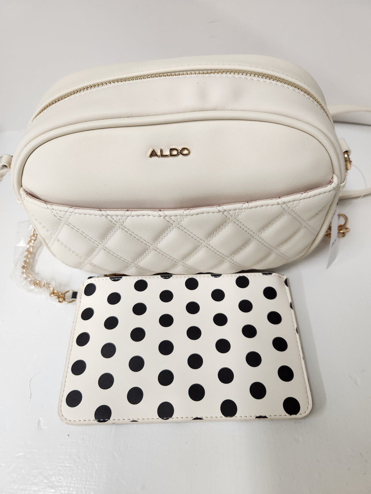 An off-white leather crossbody bag with gold hardware and the brand name 'ALDO' on the front, displayed with a matching off-white and black polka dot wallet on a white background.