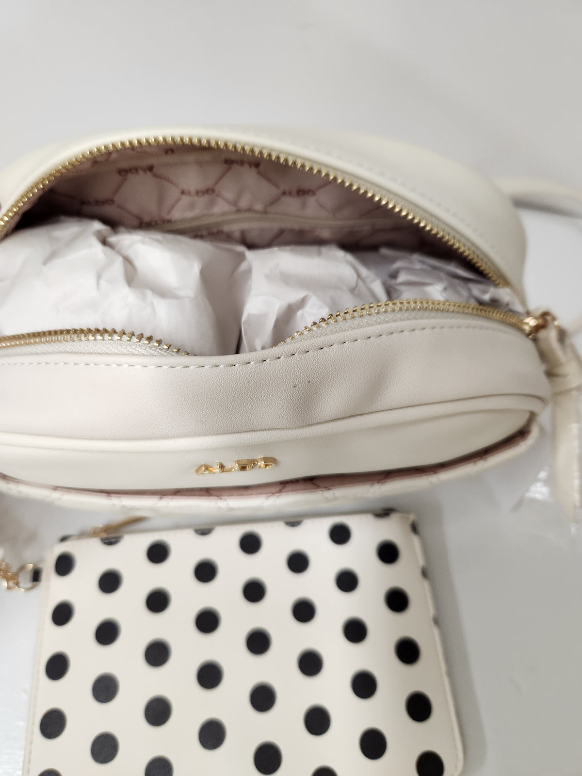 An off-white leather pouch with gold zippers and a small gold ALBE logo, shown open with tissue paper inside, alongside a matching off-white and black polka dot pouch on a white background.