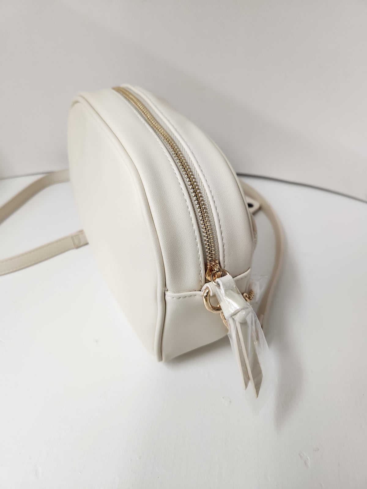 A small, off-white faux leather crossbody bag with a gold zipper and a matching off-white ribbon charm, resting on a white surface.