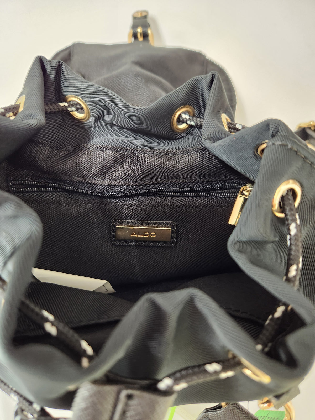 Close-up of the interior of a dark gray drawstring backpack, showing a black nylon lining, a zippered pocket, and a small metal brand plaque that reads Aldo.