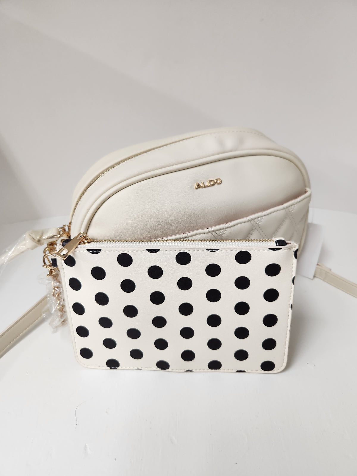 An Aldo brand cream-colored cosmetic pouch with a black polka dot patterned front zip pouch, displayed on a white surface against a white background