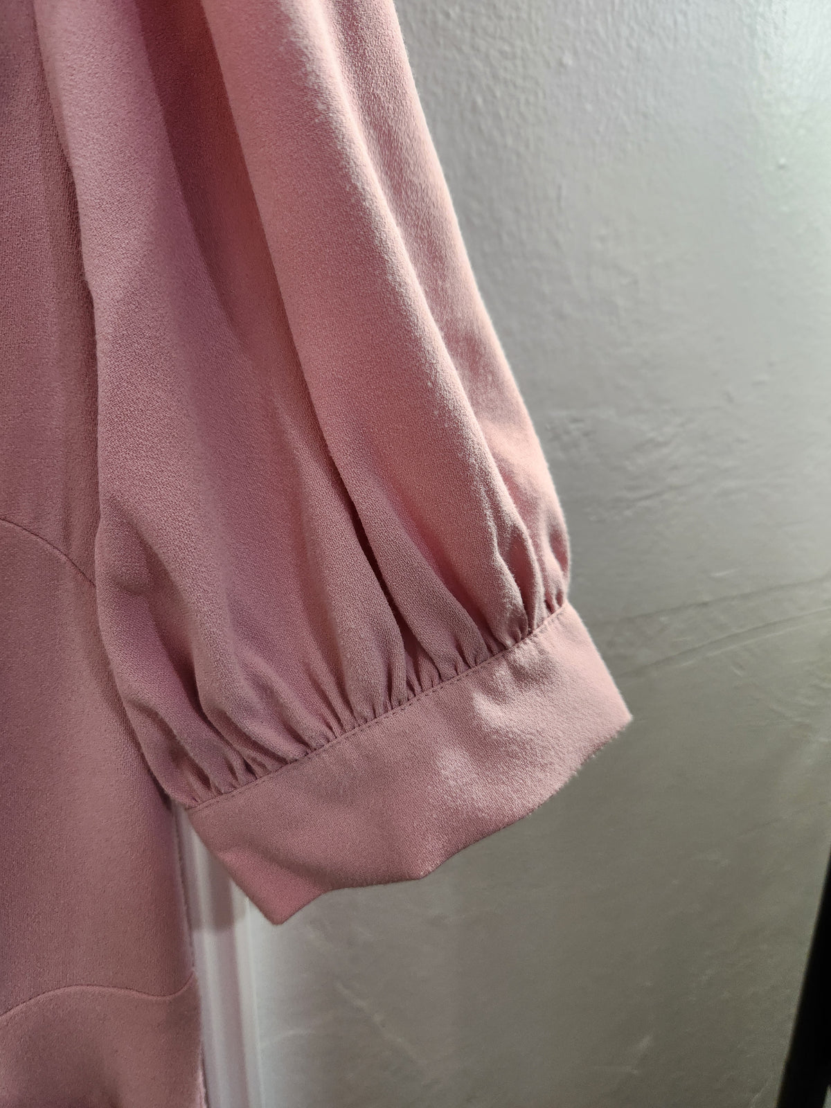 Close-up of a dusty pink garment sleeve with a gathered elastic cuff, shown against a plain light gray wall.