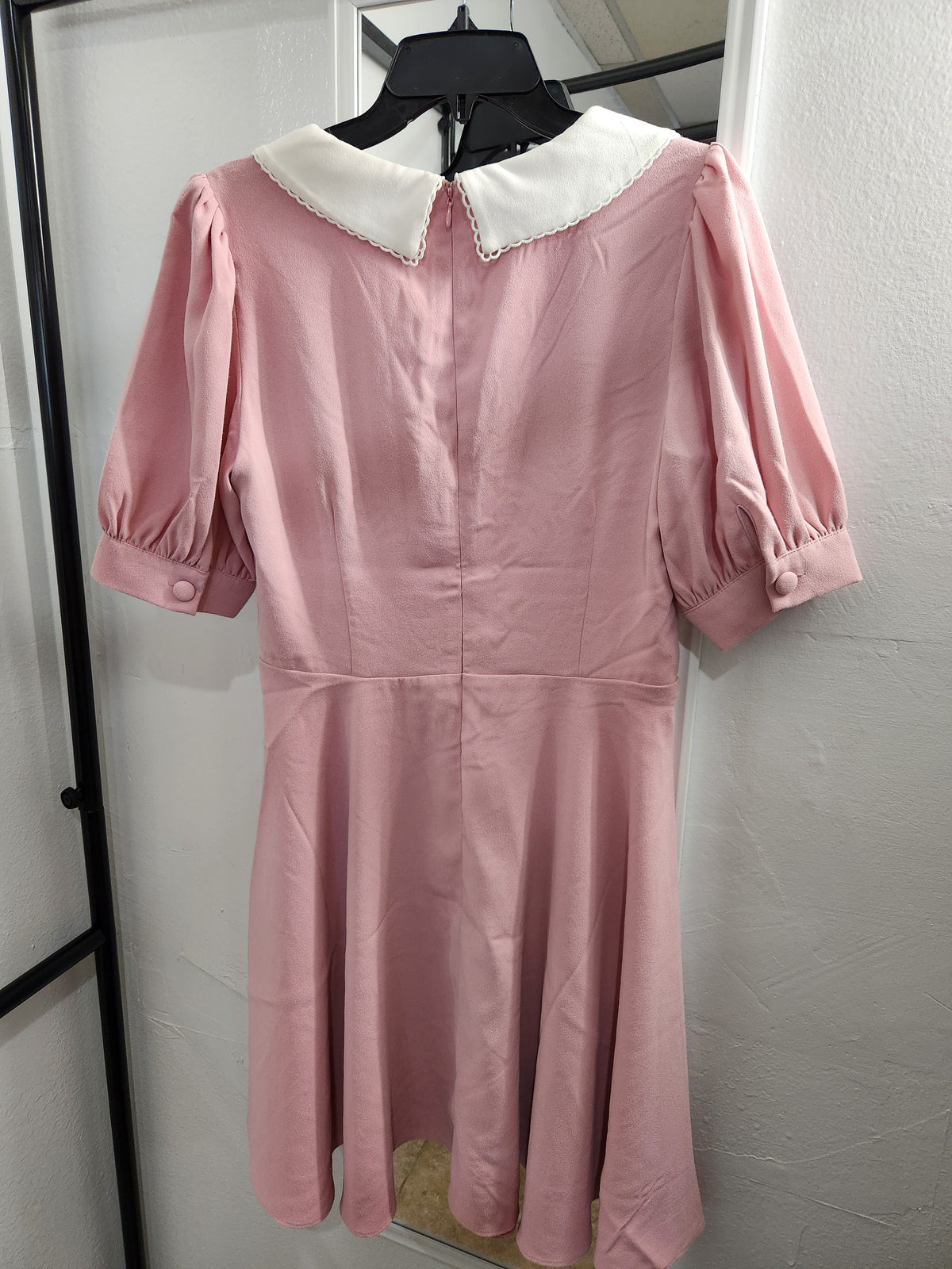 A pink short-sleeve dress with a white Peter Pan collar, hanging on a black hanger against a white wall.
