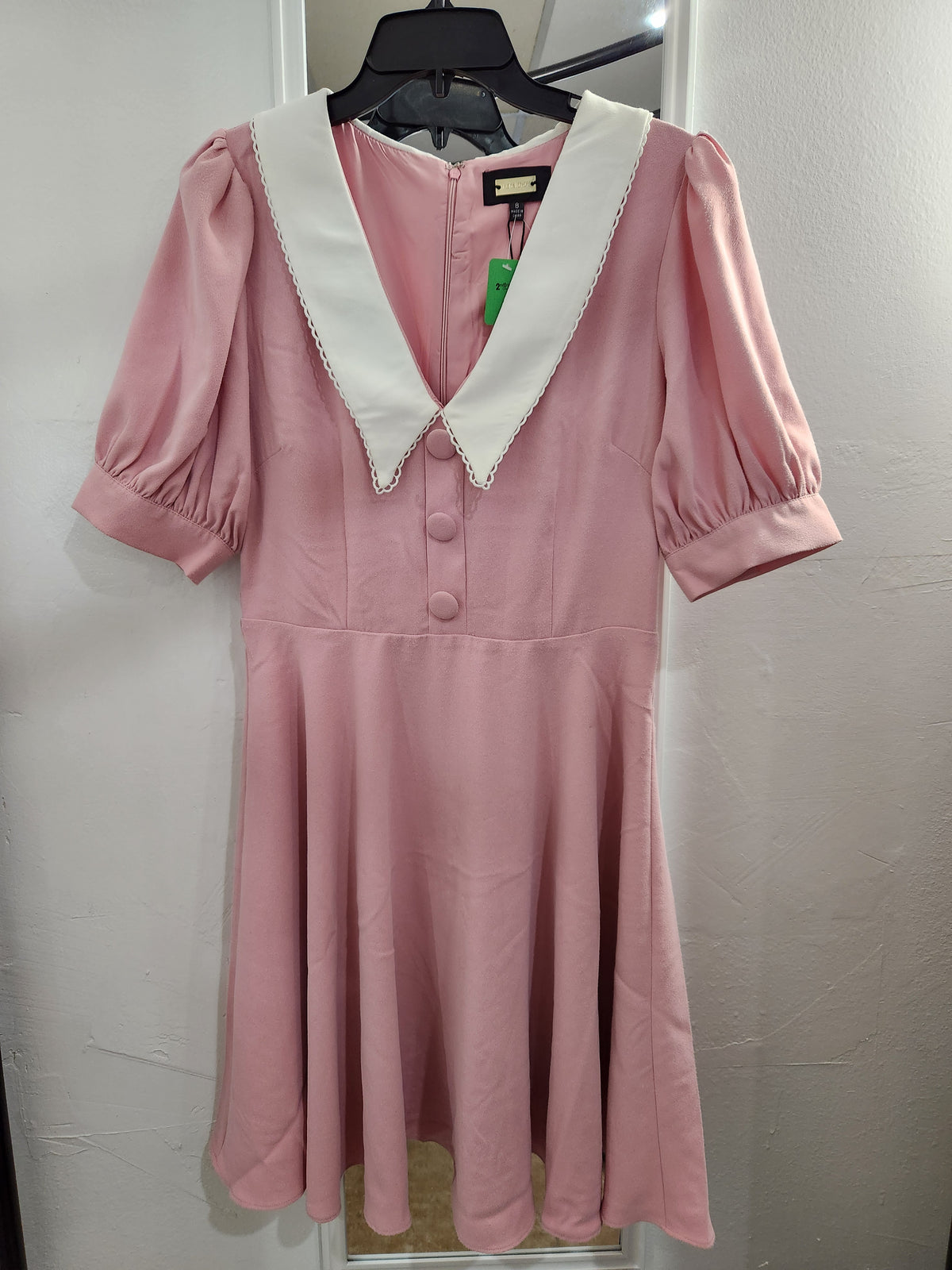 A pink short-sleeve dress with a contrasting white pointed collar and decorative trim, hanging on a black hanger against a light gray wall.