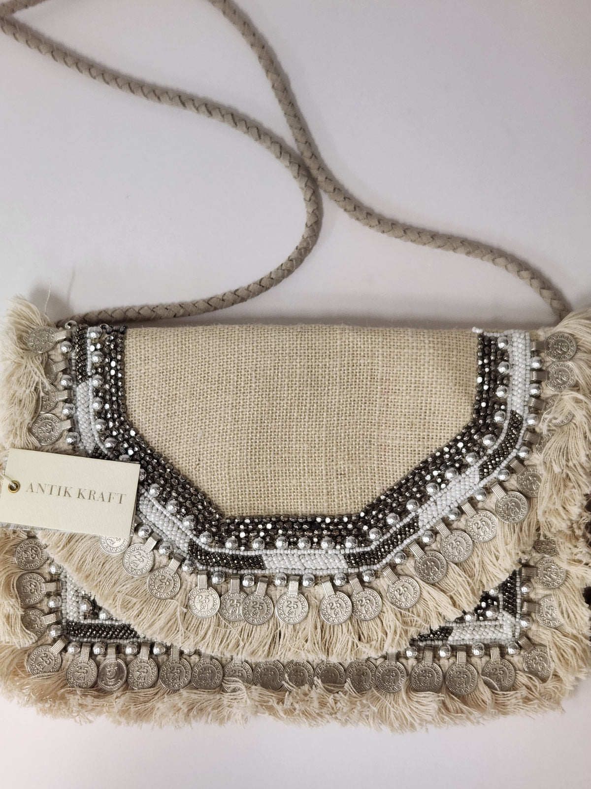 A bohemian-style clutch bag made of natural burlap with intricate silver beadwork, fringe details, and hanging silver coins, featuring a thick braided shoulder strap and a paper tag that reads 'ANTIK KRAFT'.