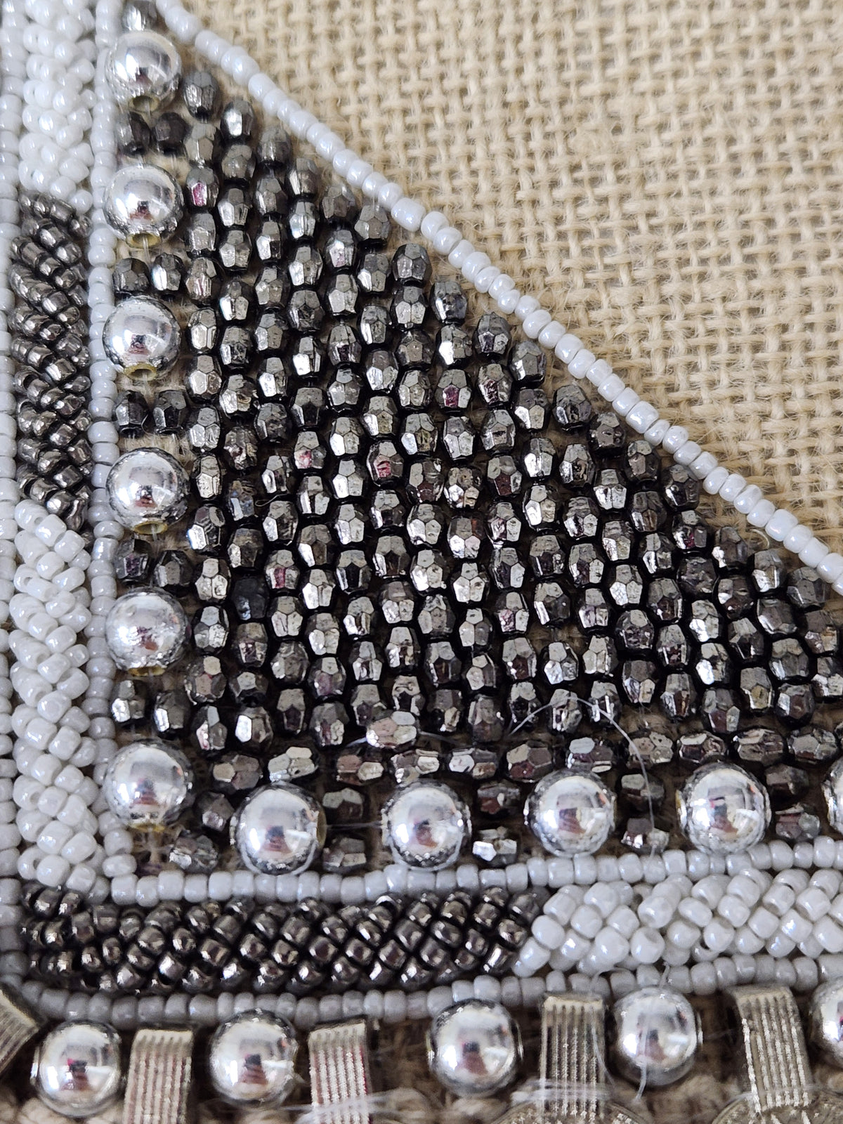 Close-up of a beaded textile corner featuring a triangular pattern of black faceted beads, silver seed beads, and white beads on a beige burlap-like background.