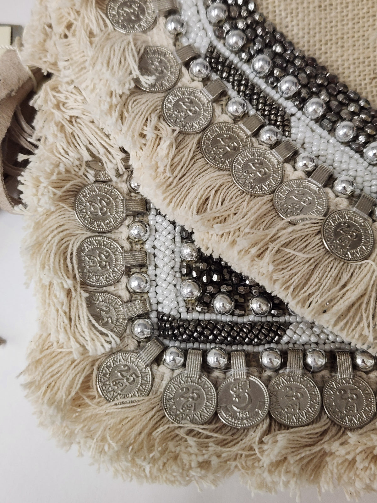 A close-up of a bohemian-style purse featuring beige fringe, silver coin embellishments, and black and white beaded geometric borders.