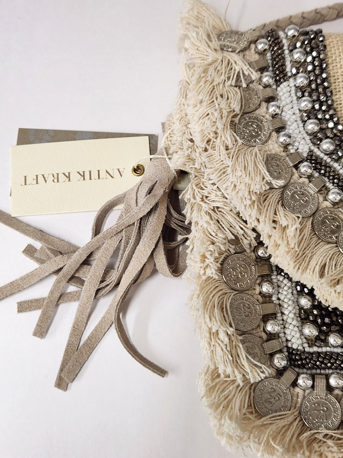 A close-up of a beige fringed textile accessory with suede-like tassels, silver coin embellishments, and black-and-silver beadwork. A paper tag reading 'ANTI KRAFT' is attached on a white background.