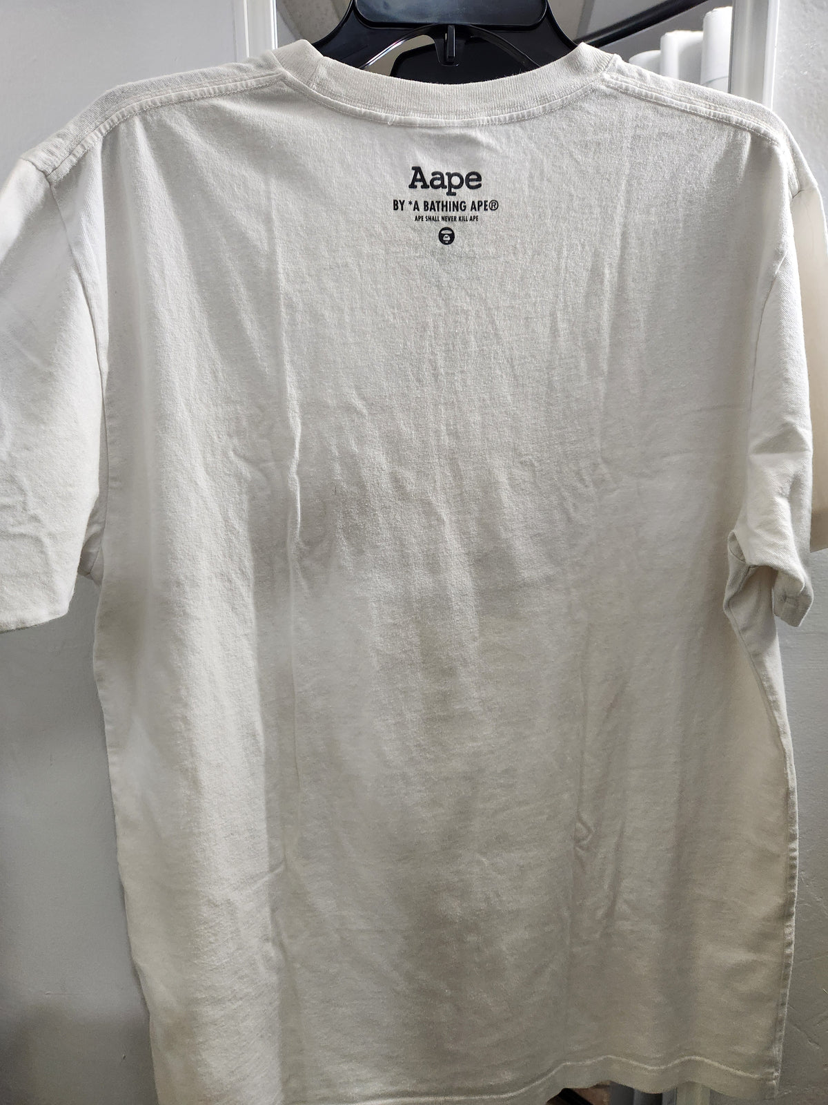 An off-white cotton t-shirt hanging on a black hanger against a light gray background. The back of the shirt features a small black printed logo for 'Aape' by Yeezy Marketing JAP at the center collar.