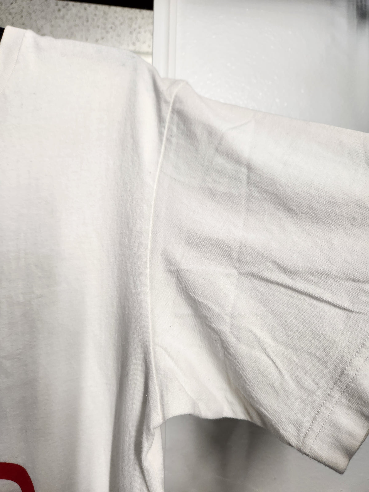 A close-up of a white short-sleeve t-shirt showing the shoulder seam and sleeve, with a small red graphic partially visible at the bottom left.