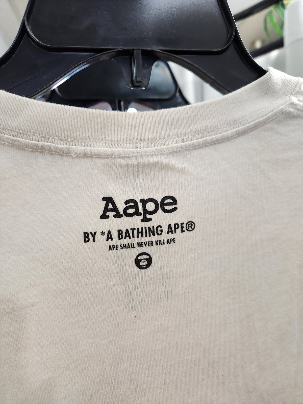 Close-up of a light beige Aape t-shirt hanging on a black hanger, showing the black printed 'Aape BY A BATHING APE' logo on the chest.