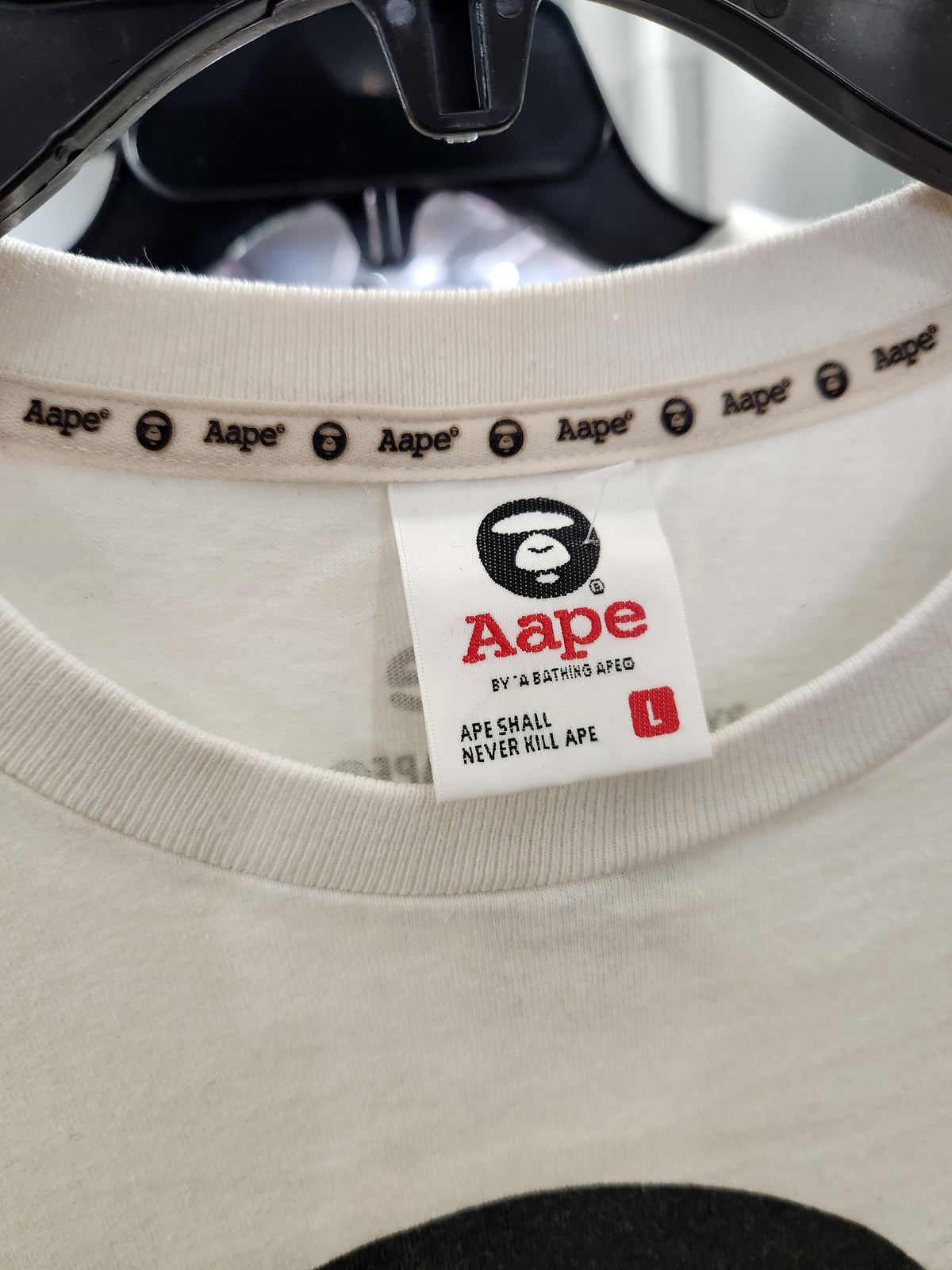 Close-up of the inner neck label of a light beige Aape t-shirt hanging on a black hanger. The label features the red and black Aape logo and text, and the inside collar is printed with the Aape brand name repeatedly.