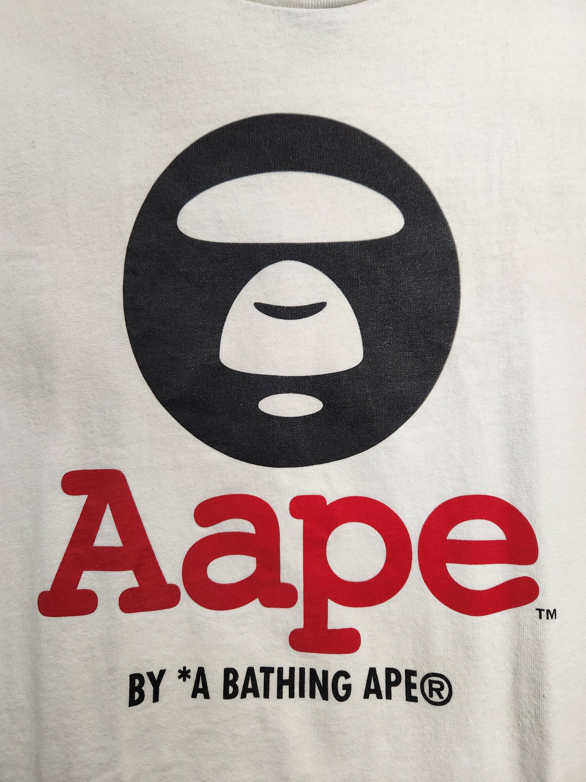 Close-up of a white graphic T-shirt featuring the black A Bathing Ape ape head logo above large red 'Aape' text, with smaller black 'BY *A BATHING APE' text below.