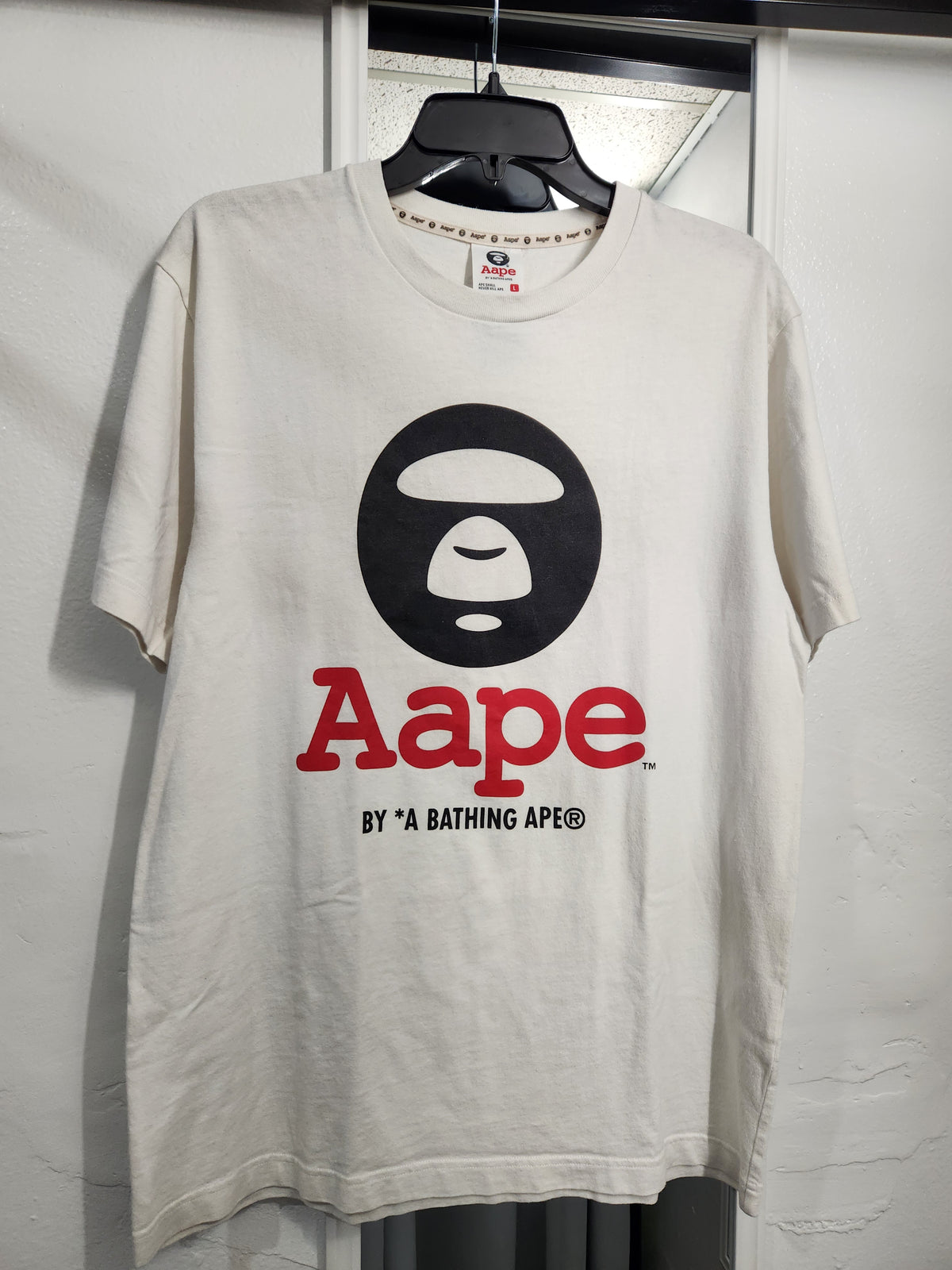 A cream-colored Aape x A Bathing Ape t-shirt hanging on a black hanger against a white wall. The shirt features the iconic black ape head logo and the word 'Aape' in red, with 'BY *A BATHING APE®' printed below in black.