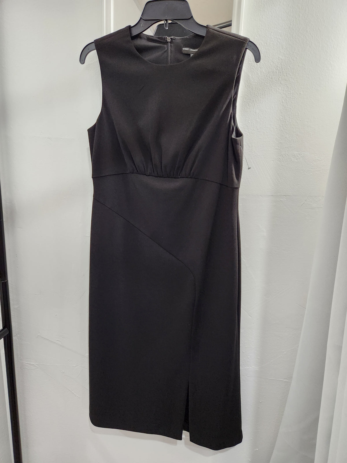 A sleeveless black knee-length dress with a gathered waist and diagonal seam detail on the skirt, hanging on a black hanger against a white curtain backdrop.