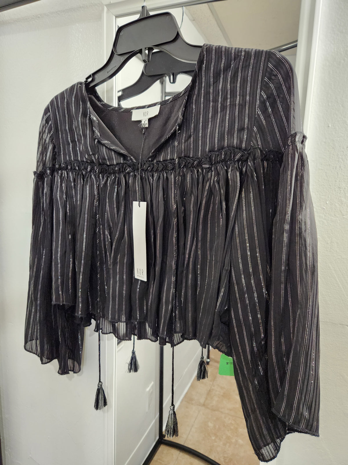 A black sheer long-sleeve blouse with vertical silver glitter stripes, a smocked neckline, and tassel ties, hanging on a black hanger.