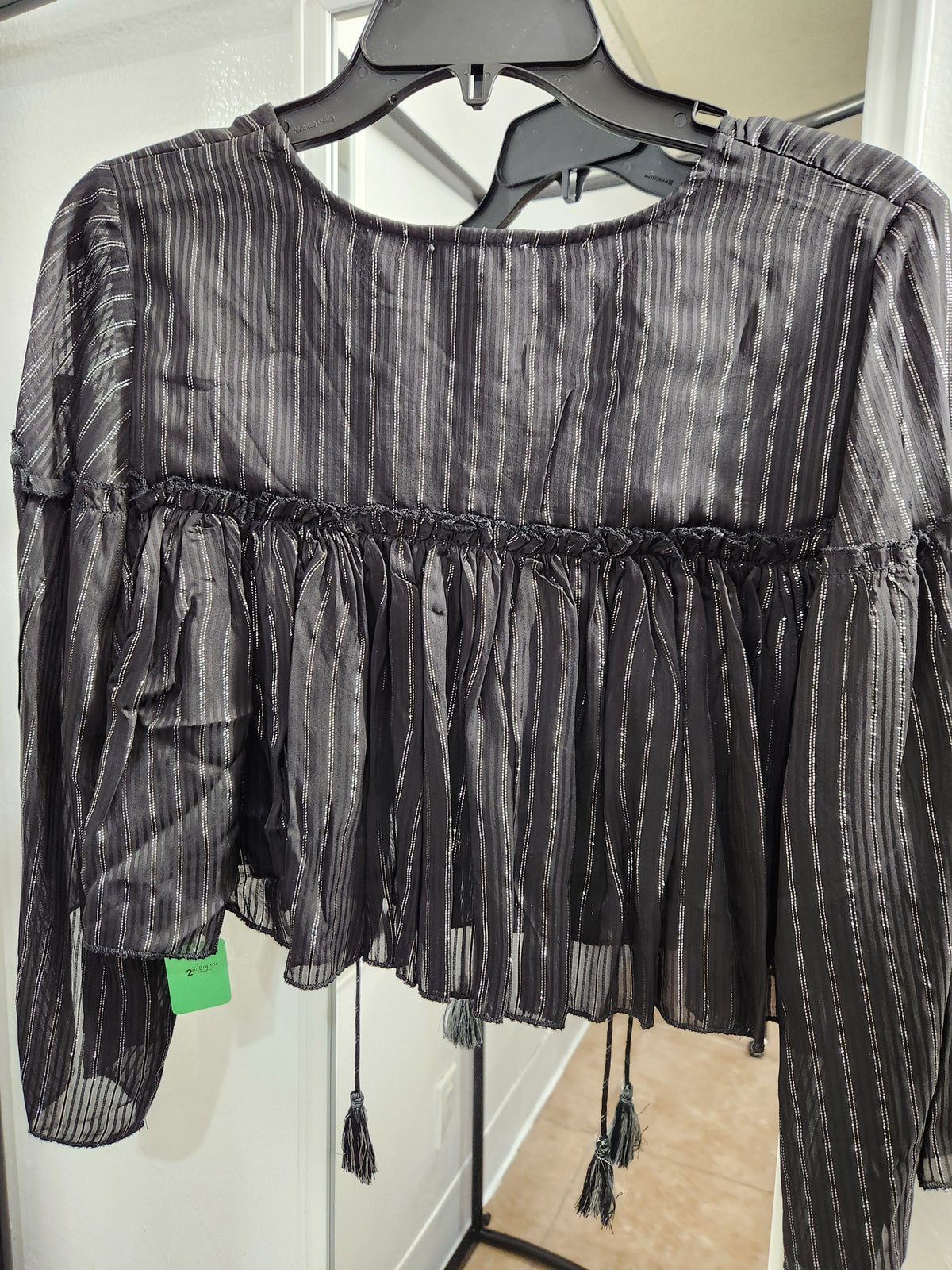 A black sheer long-sleeve blouse with vertical metallic silver stripes, a smocked bodice, and a gathered ruffled hem with tassel ties, hanging on a black hanger.