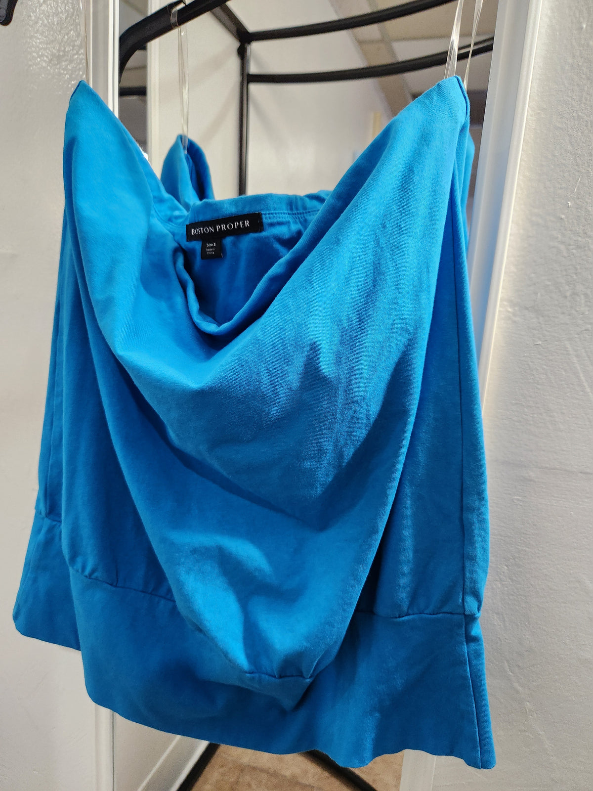 A bright blue cowl-neck top by BRON PROPER, hanging on a black clothing rack against a white wall.