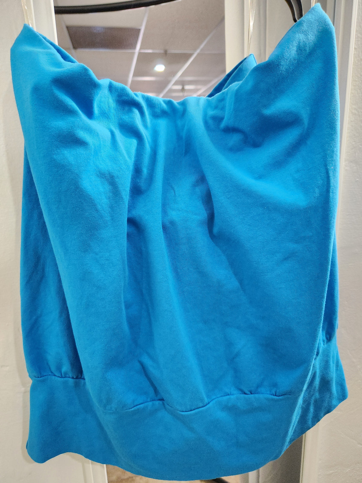 A bright blue sleeveless top or dress hanging on a black hanger in front of a white door, showing a soft, draped fabric texture.