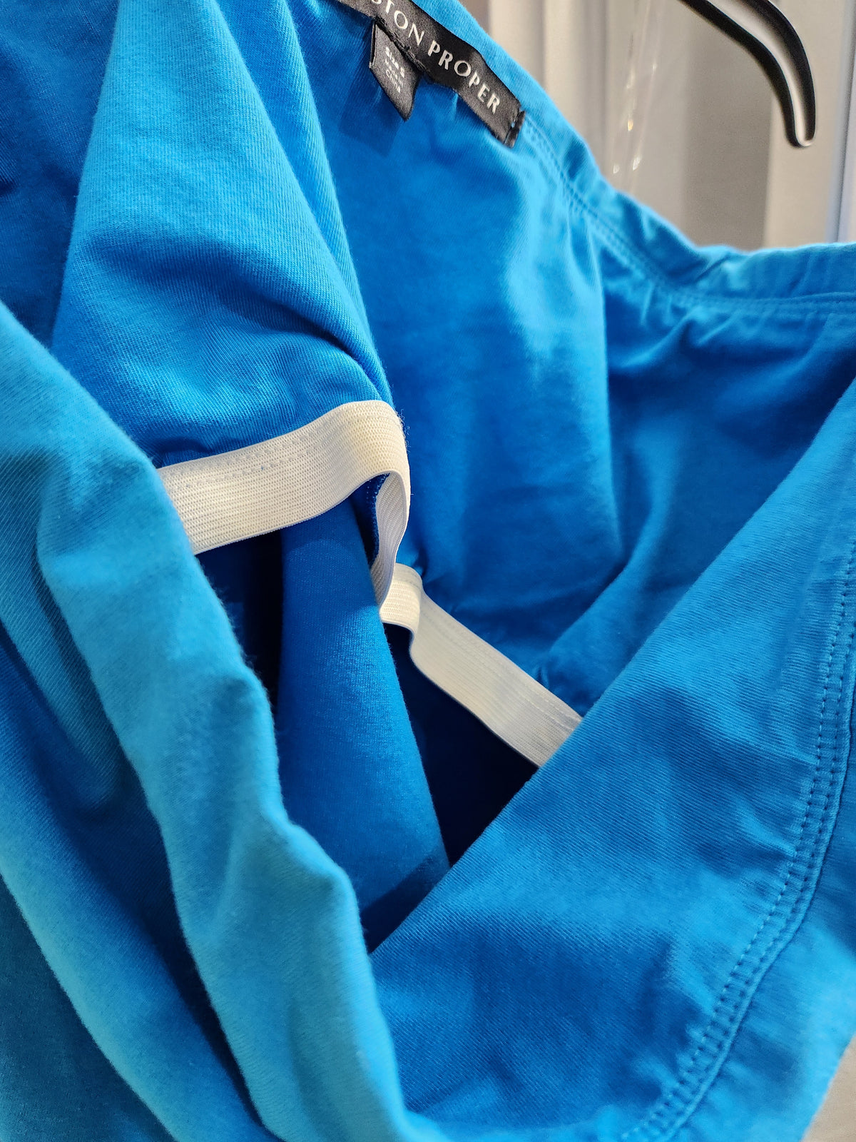 Close-up of a bright blue garment, likely a scrub top or medical uniform, with a white elastic band and a visible black neck label reading 'SAXON PROPER'.