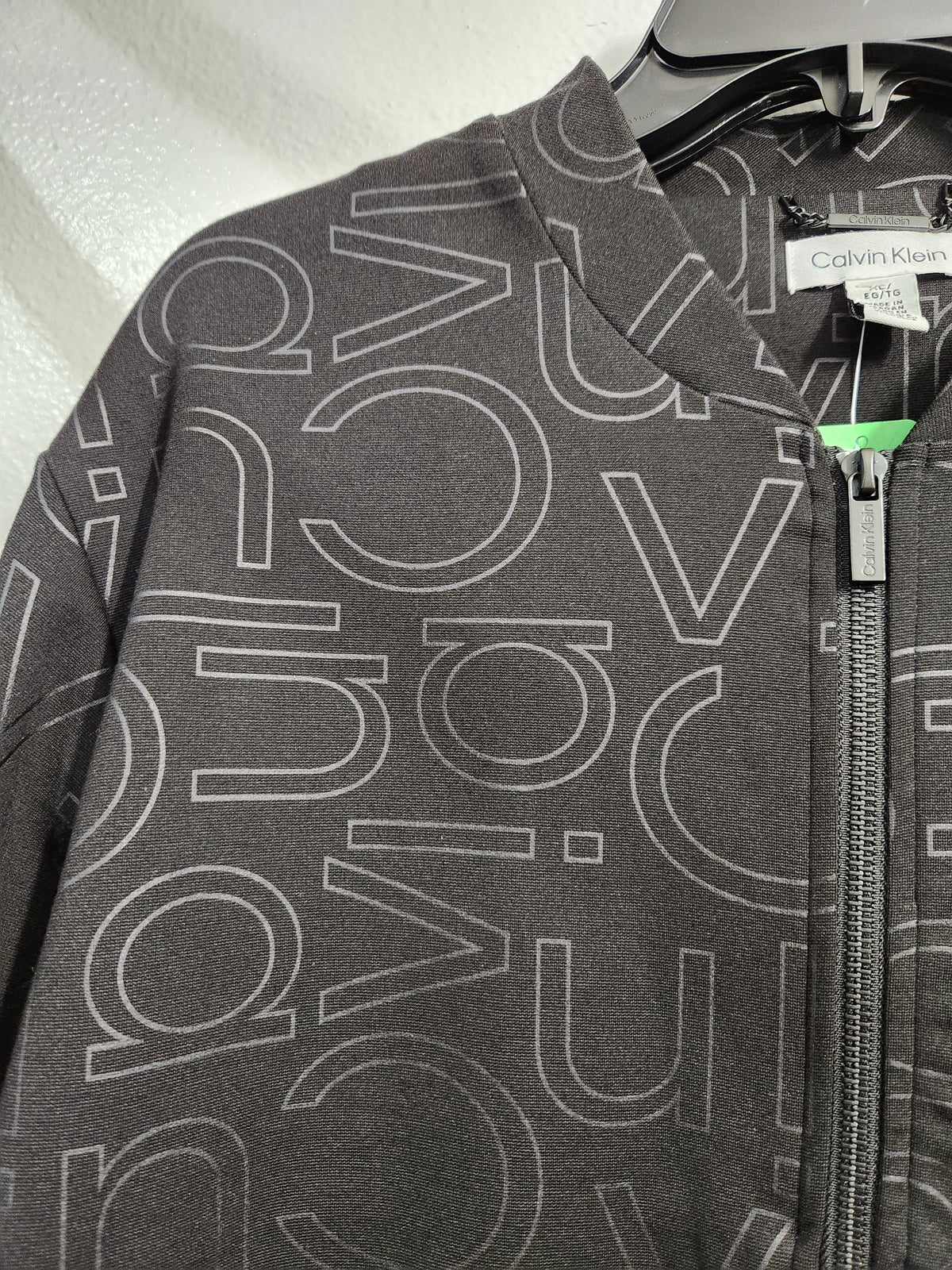 A close-up of a dark gray zip-up jacket featuring an all-over white outline of the Calvin Klein logo, hanging on a black hanger against a light textured wall.
