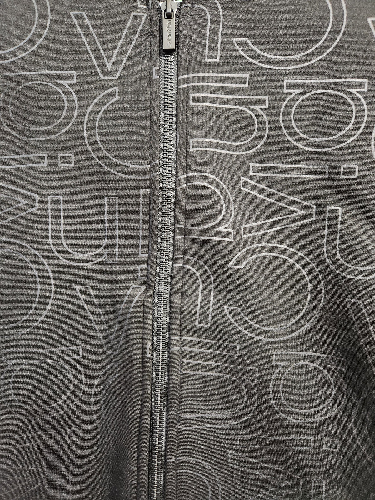 Close-up of a dark gray zip-up jacket featuring an all-over white outline pattern of stylized letters, with a silver metal zipper running down the center.