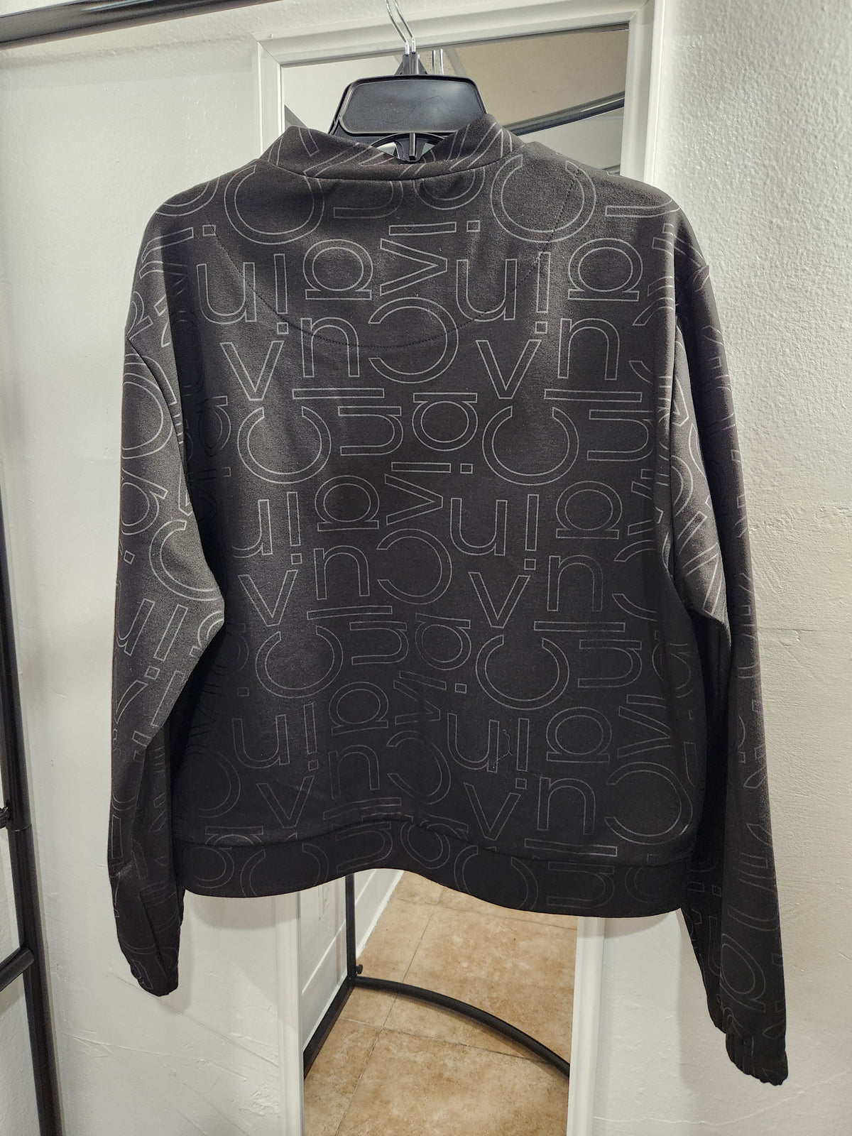 A black long-sleeve mock-neck sweatshirt with an all-over white 'Gucci' logo pattern, hanging on a black hanger in front of a white door.
