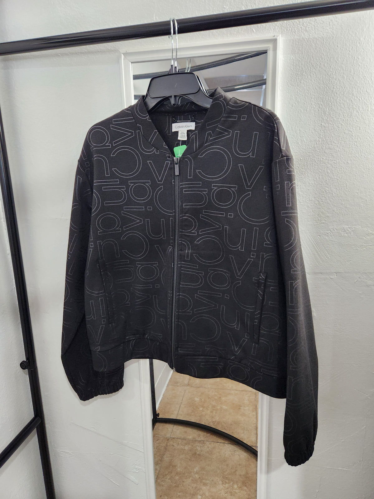 A black zip-up jacket with an all-over tonal 'DO NOT DISTURB' pattern, hanging on a black hanger on a clothing rack in front of a mirror.